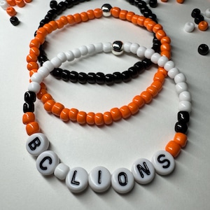 May include: Three beaded bracelets in black, orange, and white. One bracelet spells out "BC LIONS" in white letter beads. The other bracelets are solid colors with silver accent beads. Beads are scattered around the bracelets.