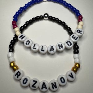 May include: Two beaded bracelets. The top bracelet is blue with a silver bead and accents of white and maroon. The bottom bracelet is black with white letter beads spelling "HOLLANDER". The bottom bracelet has gold beads and white letter beads spelling "ROZANOV".