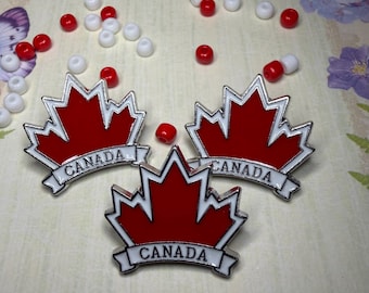 May include: Three enamel pins shaped like red maple leaves with white borders and a white banner at the bottom featuring the word CANADA in black capital letters. The pins are arranged in a triangular formation among scattered red and white beads.