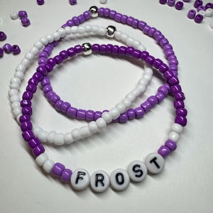 May include: Three layered beaded bracelets in shades of purple and white with the word "FROST" spelled out in white beads with black lettering.