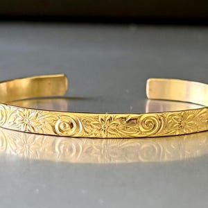 May include: A gold bangle bracelet with a floral design. The bracelet is made of a smooth, shiny metal and has a simple, elegant design.