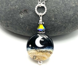 May include: A silver chain necklace with a round pendant featuring a crescent moon design on a black background with a white border. The pendant is layered with a tan and white agate-like material. A small iridescent crystal hangs above the pendant.