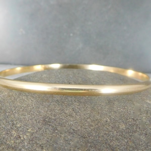 May include: A gold-colored bangle bracelet. The bracelet is a simple, smooth band with a rounded profile. The jewelry is displayed on a gray stone surface, with a blurred gray background.