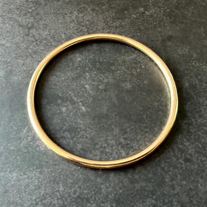 May include: A gold-colored bangle bracelet. The circular bracelet is made of a smooth, shiny metal. The bangle is a simple, elegant piece of jewelry. The bracelet is shown on a dark gray surface.