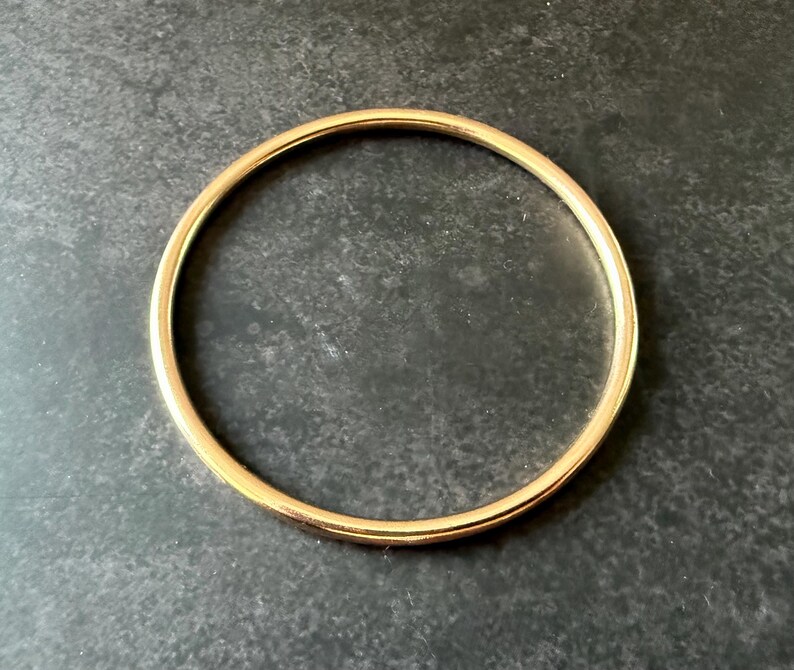 May include: A gold-colored bangle bracelet. The circular bracelet is made of a smooth, shiny metal. The bangle is a simple, elegant piece of jewelry. The bracelet is shown on a dark gray surface.