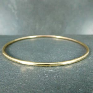 May include: A gold-colored bangle bracelet. The circular bracelet has a smooth, slightly textured surface. The jewelry is displayed on a dark gray surface. The bangle is a simple, elegant piece of jewelry.