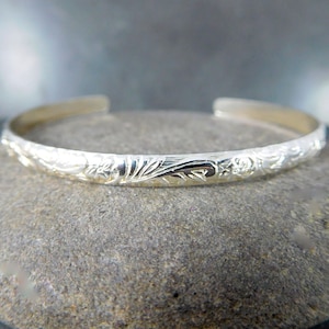 May include: A silver bangle bracelet with a floral design. The bracelet is thin and delicate, and the floral design is intricate and detailed.