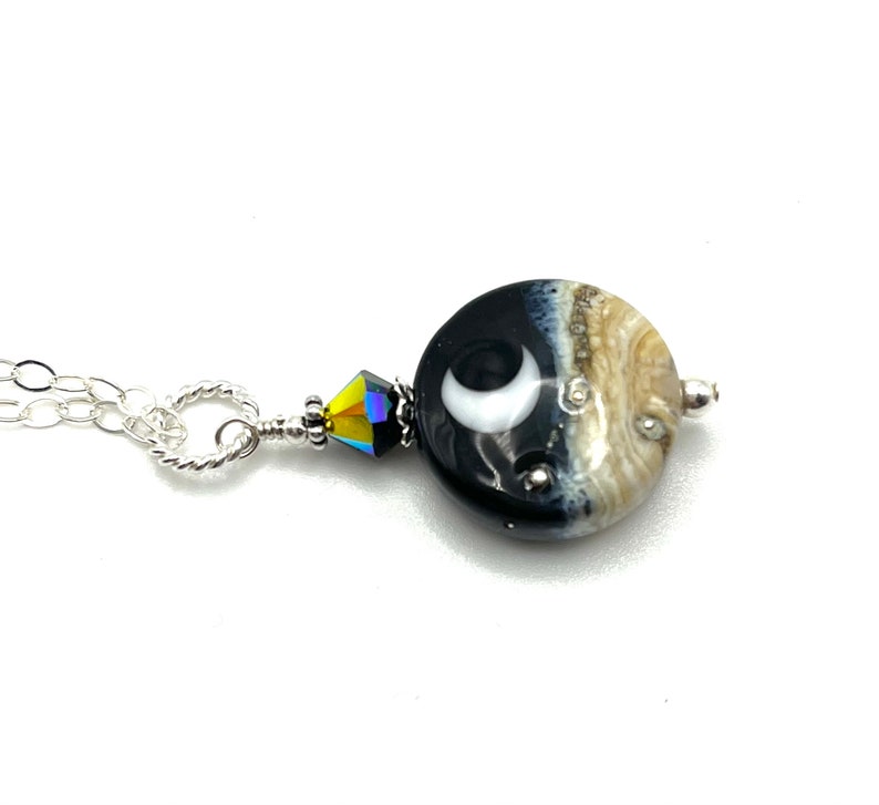 May include: A silver chain necklace with a black and white glass pendant featuring a crescent moon design. The pendant is accented with a clear crystal bead.