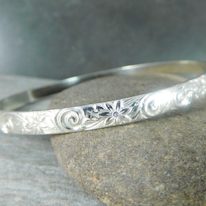 May include: A silver bangle bracelet with an intricate floral design. The bracelet is smooth and polished, with a delicate floral pattern.