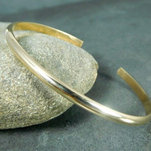 May include: A simple gold bangle bracelet, made of polished metal, is displayed on a gray rock.