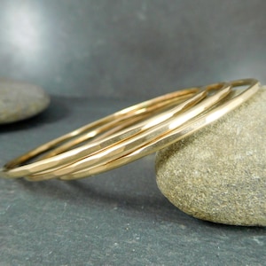 May include: Three gold-toned hammered metal bangles stacked together. The bangles are smooth and shiny, and they have a slightly irregular shape.