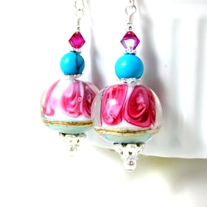 Könnte beinhalten: Ein Paar Hängeohrringe mit einer pinkfarbenen und weißen Glasperle mit Swirl-Muster, einer türkisfarbenen Perle und einer pinkfarbenen Kristallperle.