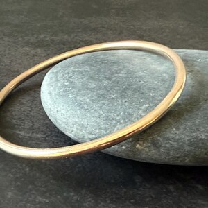 May include: A gold-colored bangle bracelet. The circular bracelet is resting on a gray stone. The bangle has a smooth, polished surface and a simple, elegant design. The background is a dark, textured surface.