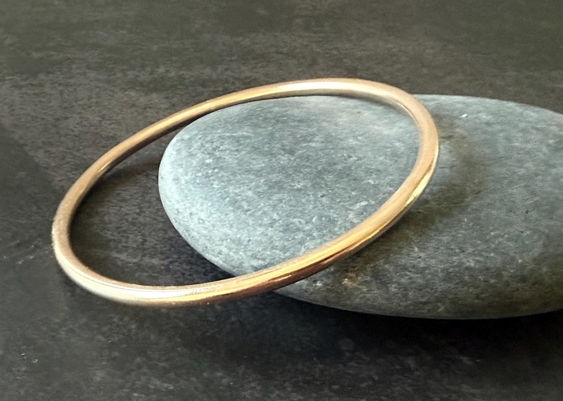 May include: A gold-colored bangle bracelet. The circular bracelet is resting on a gray stone. The bangle has a smooth, polished surface and a simple, elegant design. The background is a dark, textured surface.