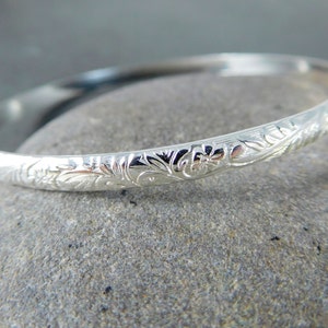 May include: A silver bangle bracelet with an intricate floral design. The bracelet is smooth and polished, and the floral pattern is raised and detailed.