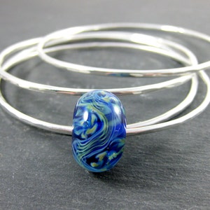May include: Three silver bangles stacked on top of each other with a blue and green glass bead in the center.