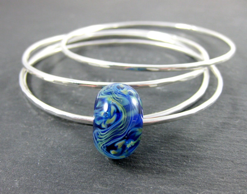 May include: Three silver bangles stacked on top of each other with a blue and green glass bead in the center.