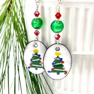 May include: A pair of dangle earrings featuring a white oval charm with a green Christmas tree with colorful lights and a star on top. The charm is attached to a silver wire with a green and red glass bead.