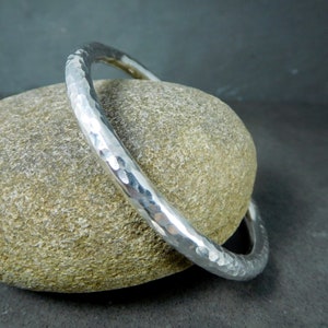 May include: A silver hammered bangle bracelet. The bracelet is thick and has a rounded shape. It is resting on a gray rock.