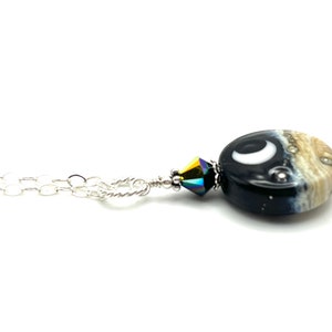 May include: A silver chain necklace with a black and white glass bead pendant featuring a crescent moon and stars. The bead is accented with a iridescent crystal.