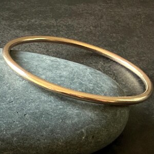 May include: A gold-colored bangle bracelet resting on a gray stone. The bracelet is a simple, circular design, with a smooth, polished surface. The background is a dark, textured surface.