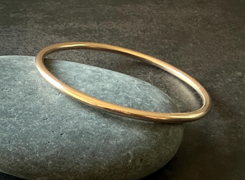 May include: A gold-colored bangle bracelet resting on a gray stone. The bracelet is a simple, circular design, with a smooth, polished surface. The background is a dark, textured surface.