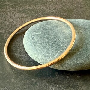 May include: A simple, gold-colored bangle bracelet. The bracelet is circular and appears to be made of metal. The bangle is resting on a gray stone, with a dark background.