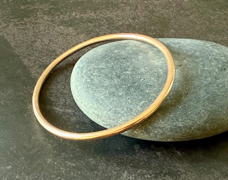 May include: A simple, gold-colored bangle bracelet. The bracelet is circular and appears to be made of metal. The bangle is resting on a gray stone, with a dark background.