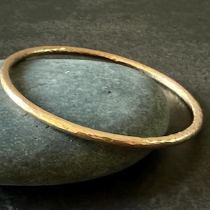 May include: A hammered gold bangle bracelet rests on a gray stone. The bracelet has a textured surface and a circular shape. The gold color is warm and the bracelet appears to be handmade.