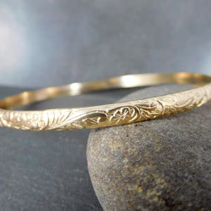 May include: A gold bangle bracelet with an intricate floral design. The bracelet is smooth and shiny, and the floral pattern is raised and detailed.
