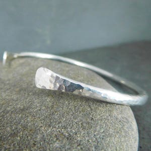 May include: A silver hammered bangle bracelet with a rounded, hammered end. The bracelet is open and has a simple, minimalist design.