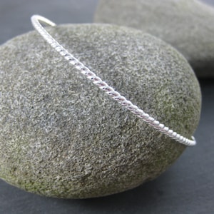 May include: A silver bangle bracelet with a twisted rope design. The bracelet is on a gray stone background.
