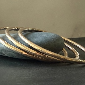 May include: Three hammered gold bangles rest on a gray stone, set against a dark background. The bangles have a textured surface and a warm, metallic sheen. The jewelry is simple and elegant.