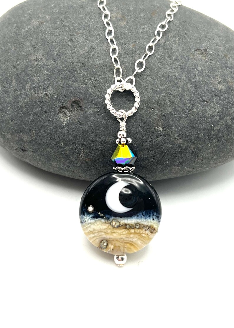 May include: A silver chain necklace with a round pendant featuring a crescent moon design on a black background with a white border. The pendant is layered with a tan and white agate-like material. A small iridescent crystal hangs above the pendant.