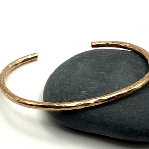May include: A hammered gold-tone cuff bracelet with a textured surface. The open-ended bracelet is resting on a dark gray stone against a white background. The jewelry piece is a simple, elegant design.