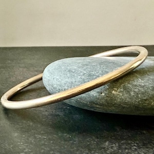 May include: A gold-colored bangle bracelet resting on a gray stone. The bracelet has a smooth, rounded design. The stone is oval-shaped and has a textured surface. The background is a neutral, dark gray.