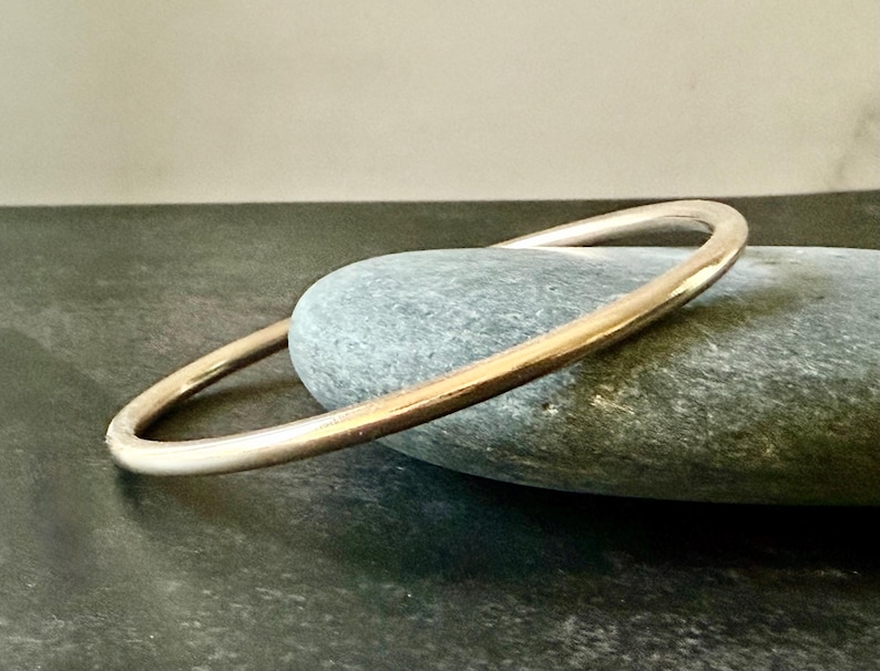 May include: A gold-colored bangle bracelet resting on a gray stone. The bracelet has a smooth, rounded design. The stone is oval-shaped and has a textured surface. The background is a neutral, dark gray.