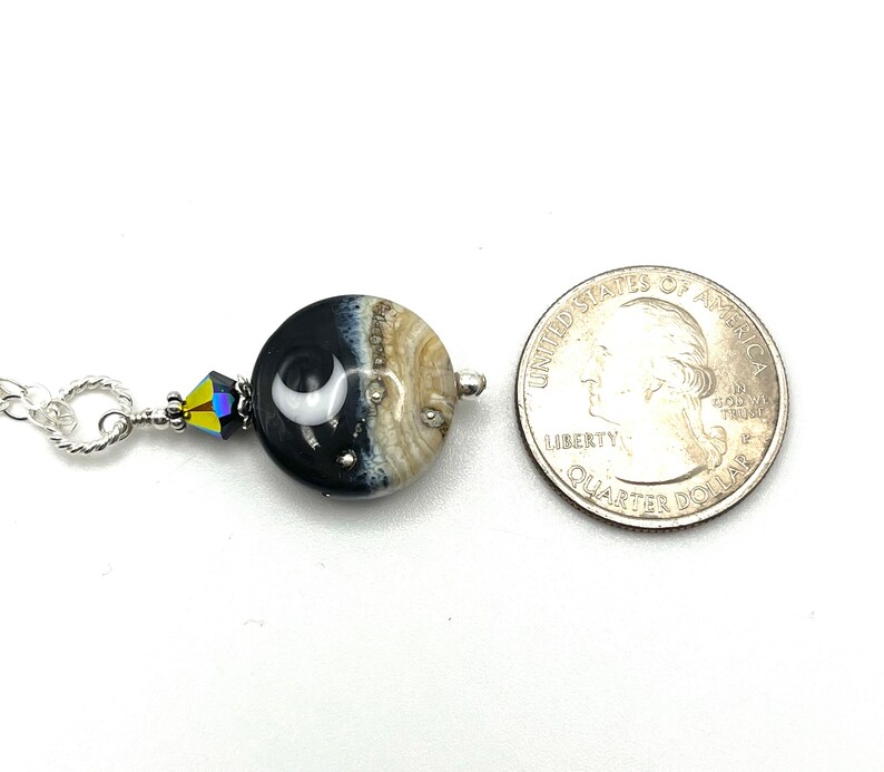 May include: A silver chain necklace with a black and white glass bead featuring a crescent moon design. The bead is accented with a clear crystal. The necklace is shown next to a US quarter for size comparison.
