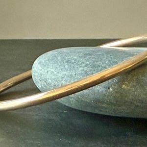 May include: A gold-colored bangle bracelet resting on a smooth, gray-green stone. The bracelet has a simple, rounded design. The stone is oval-shaped and sits on a dark surface. The lighting highlights the bracelet's metallic sheen.