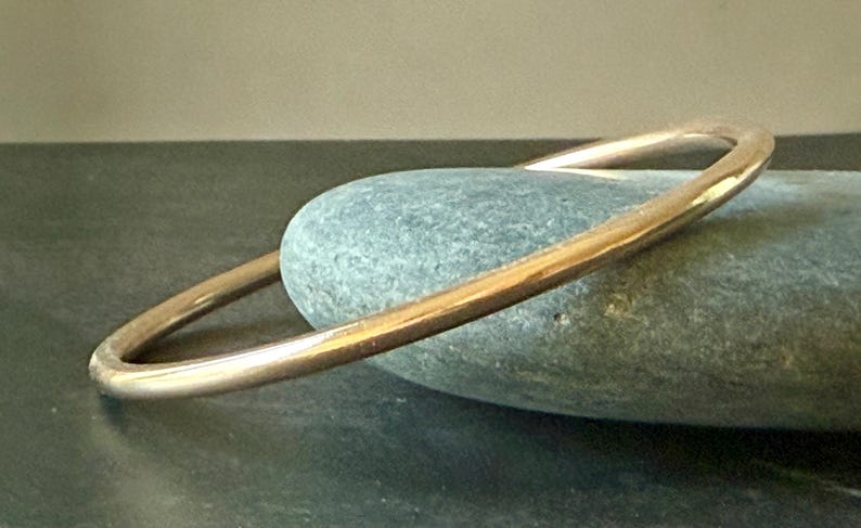 May include: A gold-colored bangle bracelet resting on a smooth, gray-green stone. The bracelet has a simple, rounded design. The stone is oval-shaped and sits on a dark surface. The lighting highlights the bracelet's metallic sheen.