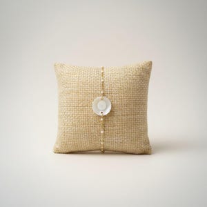 Peut inclure: Un bracelet délicat en chaîne dorée avec un pendentif circulaire blanc et un centre en forme de perle. Le bracelet est présenté sur un petit coussin en toile de jute beige. Le design est simple et élégant, avec un fond neutre.