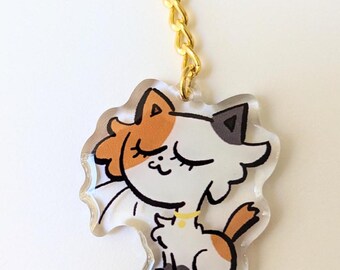 Puede incluir: Un llavero transparente con forma de gato de acrílico con una cadena dorada. La ilustración del gato presenta marcas naranjas, blancas y negras, con los ojos cerrados y un collar amarillo. El llavero está diseñado con un estilo de dibujos animados.