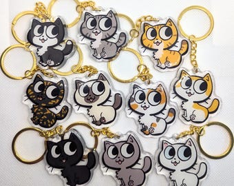 Puede incluir: Una colección de llaveros con forma de gato con anillos dorados. Los llaveros presentan varios diseños de gatos en negro, gris, naranja y blanco, con ojos grandes y caras sonrientes. Los llaveros están hechos de acrílico transparente.