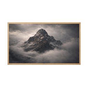 Puede incluir: Impresión enmarcada de un paisaje con una majestuosa cima montañosa envuelta en dramáticas nubes. La montaña es gris y blanca, con nieve en sus laderas superiores. El marco es de color marrón claro.