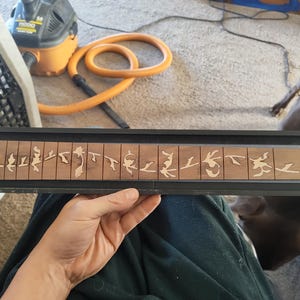 Op de afbeelding: Een houten gitaar toets met ingelegde decoratieve ontwerpen. De toets wordt in een zwart frame gehouden. De ontwerpen zijn lichtgekleurd en lijken uitgesneden vormen te zijn. De toets heeft een warme bruine kleur.