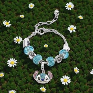 K&ouml;nnte beinhalten: Silbernes Charm-Armband mit blauen und silbernen Perlen und einem Stitch-Charm. Das Armband befindet sich auf einem gr&uuml;nen Hintergrund mit wei&szlig;en G&auml;nsebl&uuml;mchen. Der Charm ist eine Zeichentrickfigur mit blauen und rosa Akzenten.