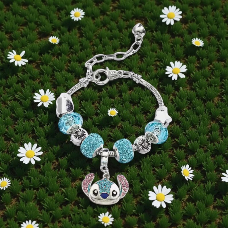 K&ouml;nnte beinhalten: Silbernes Charm-Armband mit blauen und silbernen Perlen und einem Stitch-Charm. Das Armband befindet sich auf einem gr&uuml;nen Hintergrund mit wei&szlig;en G&auml;nsebl&uuml;mchen. Der Charm ist eine Zeichentrickfigur mit blauen und rosa Akzenten.