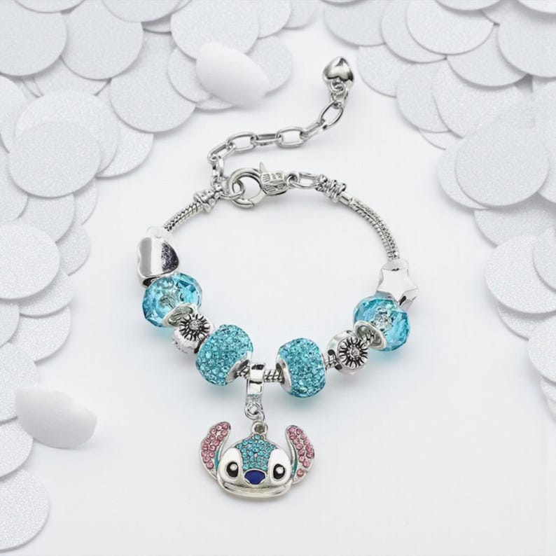K&ouml;nnte beinhalten: Silbernes Charm-Armband mit blauen und silbernen Perlen und einem Stitch-Anh&auml;nger. Der Anh&auml;nger zeigt eine Zeichentrickfigur mit blauen, rosa und silbernen Akzenten. Das Armband hat einen Kettenverschluss.