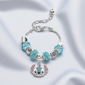 K&ouml;nnte beinhalten: Silberfarbenes Charm-Armband mit blauen Kristallperlen und einem Stitch-Anh&auml;nger. Der Anh&auml;nger hat blaue und rosa Akzente. Das Armband hat einen Karabinerverschluss und einen herzf&ouml;rmigen Anh&auml;nger. L&auml;nge ca. 18 cm.