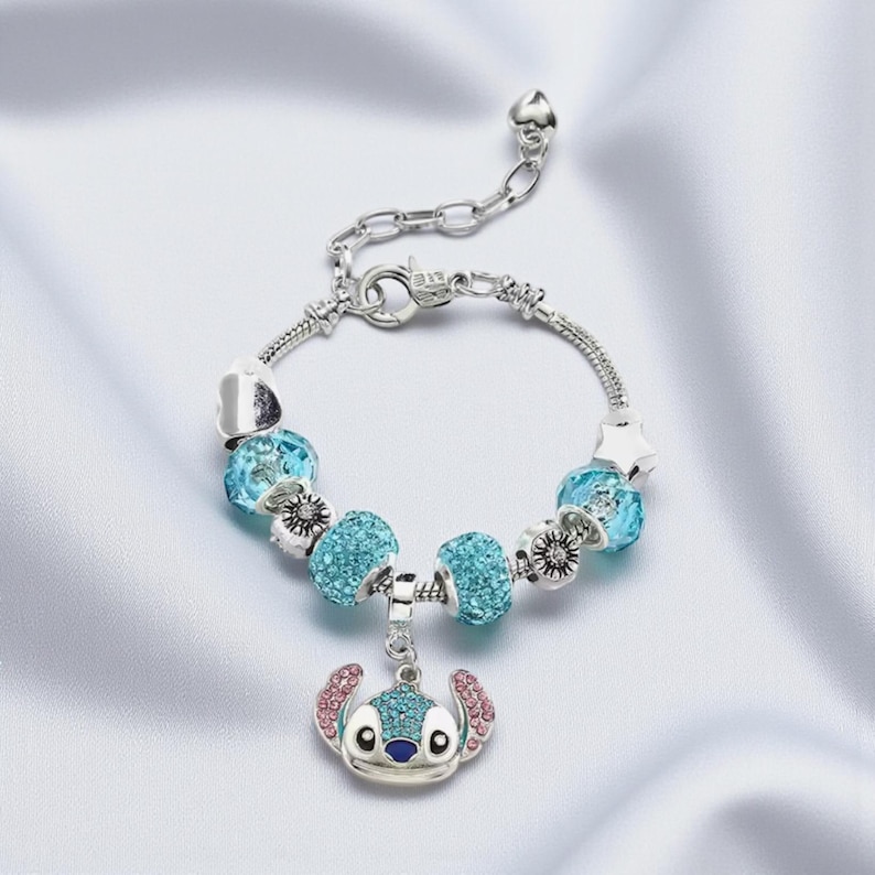 K&ouml;nnte beinhalten: Silberfarbenes Charm-Armband mit blauen Kristallperlen und einem Stitch-Anh&auml;nger. Der Anh&auml;nger hat blaue und rosa Akzente. Das Armband hat einen Karabinerverschluss und einen herzf&ouml;rmigen Anh&auml;nger. L&auml;nge ca. 18 cm.
